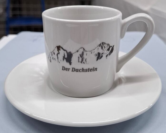 White porcelain espresso cup with black Dachstein motif and matching saucer on a light-coloured table | © Planai-Hochwurzen-Bahnen Gesellschaft m.b.H.