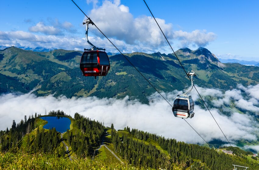 Two cable cars over Dorfgastein with forest, pond and clouds below, set against an alpine mountain backdrop. | © Dorfgasteiner Bergbahnen AG