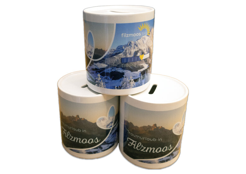 Three cylindrical Filzmoos money boxes with mountain panorama and coin slot on a light background. | © Bergbahnen Filzmoos GmbH