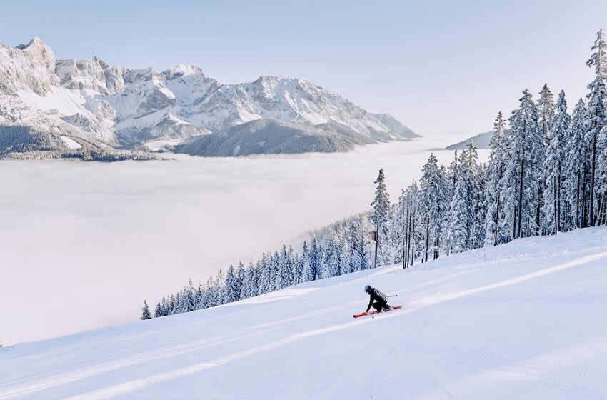 A skier descends a groomed slope with snowy mountains and a sea of fog in the background. | © Bergbahnen Filzmoos GmbH