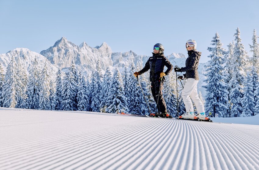 Two skiers stand on ribbed slope with snowy trees and Dachstein massif in the background. | © Bergbahnen Filzmoos GmbH