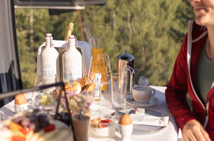 Woman sitting at laid table with breakfast and orange juice inside mountain gondola above green forest slope | © Großarler Bergbahnen GmbH & Co KG