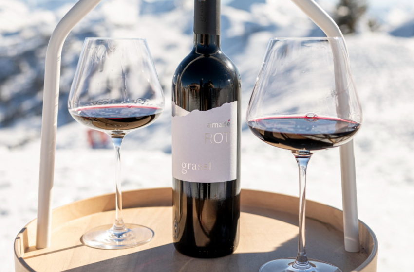 A wine bottle stands between two filled wine glasses on a small table in the snow with alpine mountains behind | © Großarler Bergbahnen GmbH & Co KG