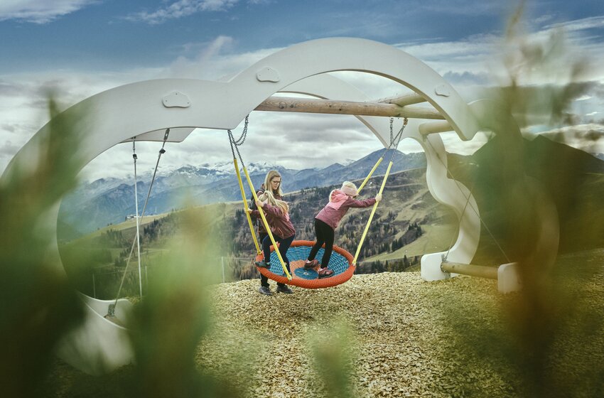 Child on a large net swing with mom, scenic mountain backdrop and modern playground structure in nature. | © Grossarler Bergbahnen GmbH & Co KG