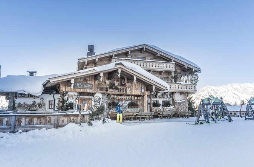 Snow-covered Abergalm with skier, skis and wooden benches before snow-covered mountains under a clear blue sky | © Creatina Werbedesign & Photography