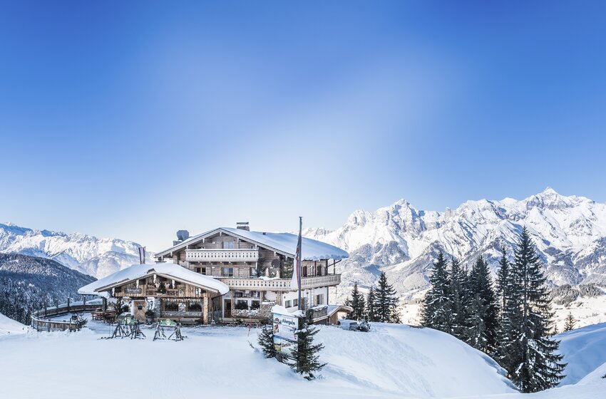 Snow-covered mountain hut with terrace and fir trees before a wide panorama of snow-covered Alpine peaks | © Creatina Werbedesign & Photography