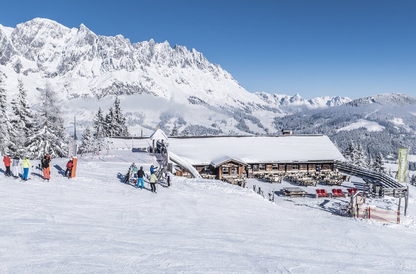Karbachalm with terrace beside a wide ski slope, surrounded by skiers and ski racks below snow-covered mountains | © Creatina Werbedesign & Photography
