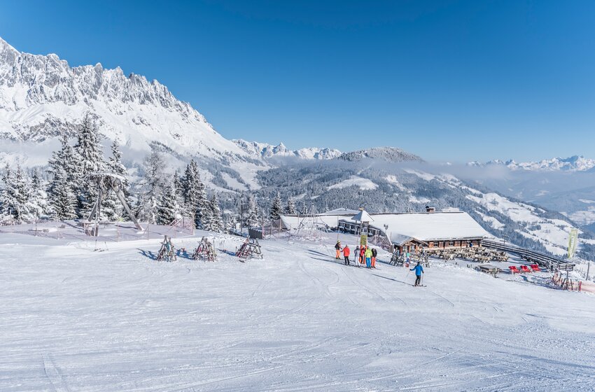 Karbachalm beside a wide ski slope with skiers and ski racks, backed by snow-covered mountains under a blue sky | © Creatina Werbedesign & Photography