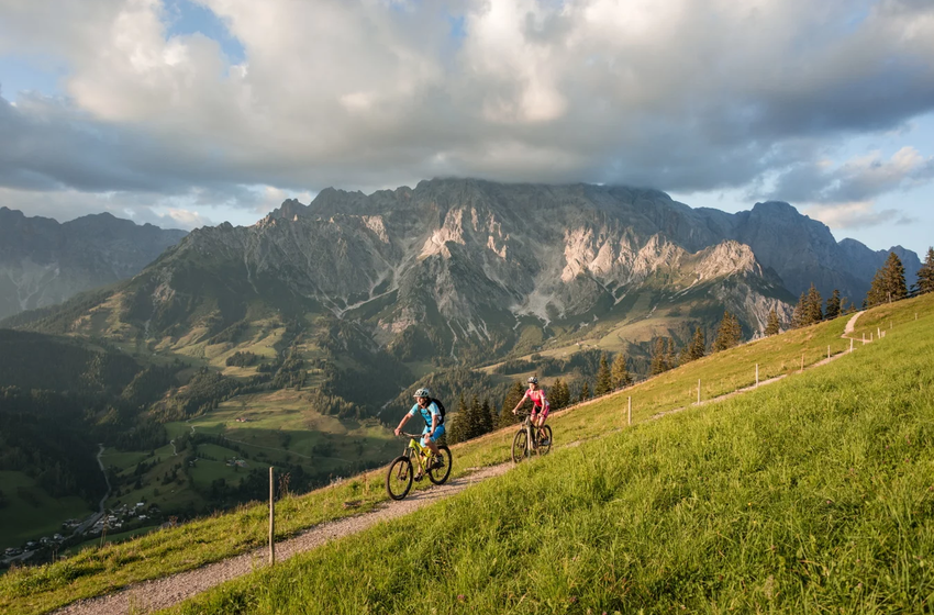 Two mountain bikers ride a narrow alpine trail at Hochkönig, surrounded by green meadows and dramatic peaks under cloudy sky | © Hochkönig Bergbahnen GmbH