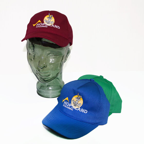 Three kids’ caps with Hopsi embroidery and 'Hopsiland Schladming' text in red, blue and green, one on glass model | © Planai-Hochwurzen-Bahnen GmbH