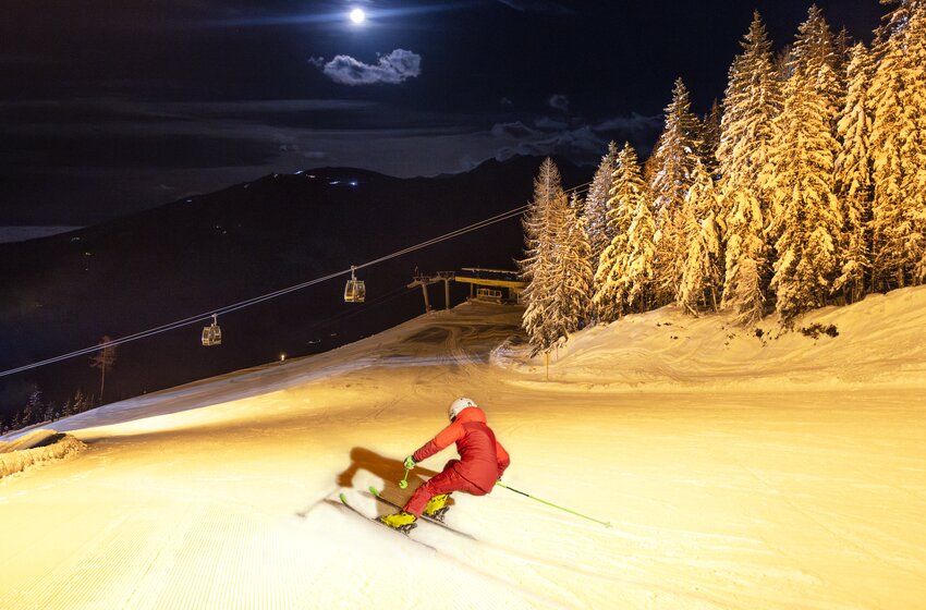Skier in red suit descends illuminated slope at night, surrounded by snowy trees and gondolas under a bright full moon. | © Planai-Hochwurzen-Bahnen Gesellschaft m.b.H.