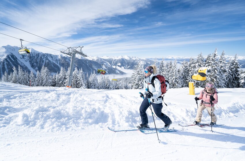 Two ski tourers hike up snowy Planai slope in sunshine with panoramic mountain views in the background.