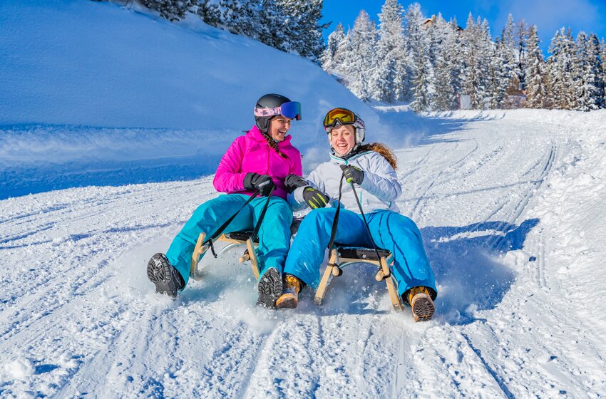 Two smiling women toboggan side by side down a snowy slope with snow spraying up around them. | © Planai-Hochwurzen-Bahnen Gesellschaft m.b.H.