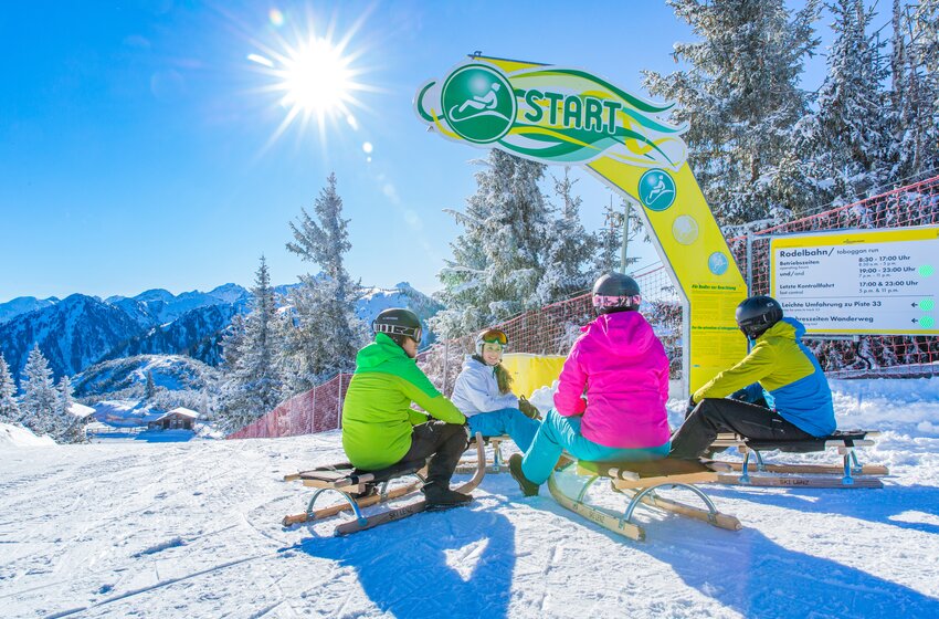 Four tobogganers sit at a sunny toboggan run start line with snowy mountain views in the background. | © Planai-Hochwurzen-Bahnen Gesellschaft m.b.H.