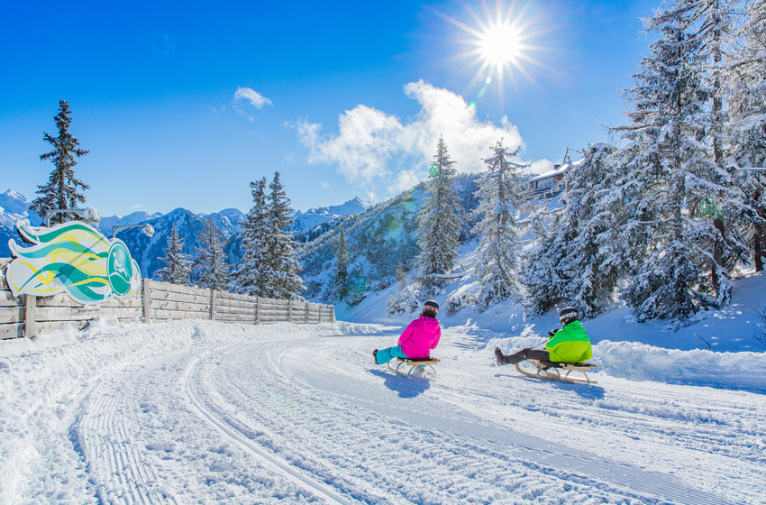 Two tobogganers ride through a sunny snowy curve with mountain and forest backdrop in brilliant winter light. | © Planai-Hochwurzen-Bahnen Gesellschaft m.b.H.