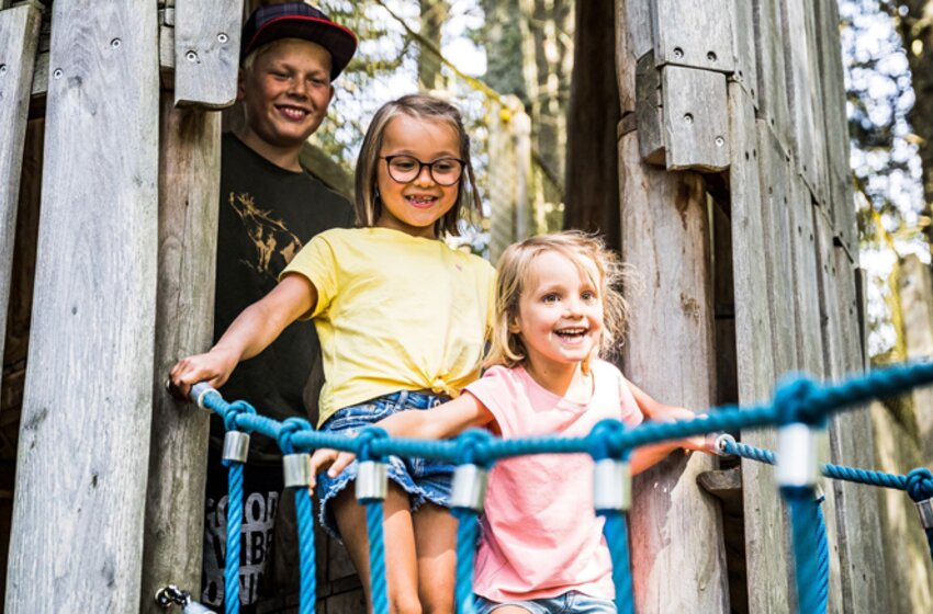Two girls happily cross a rope bridge in the forest, with a smiling teenager standing behind them at a wooden play tower. | © Snow Space Salzburg Bergbahnen