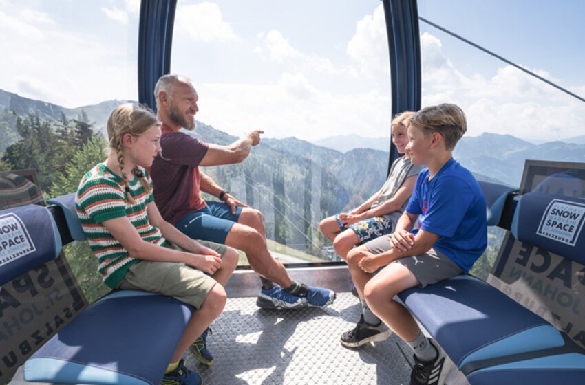 An adult sits with three children in the Flying Mozart gondola, looking out at the sunny mountain scenery outside. | © Snow Space Salzburg Bergbahnen