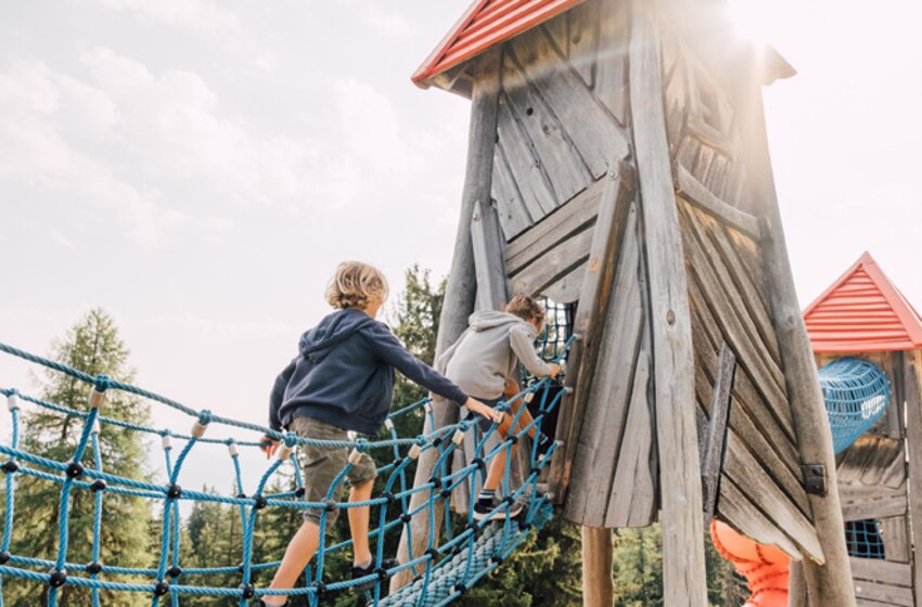 Two children climb a blue rope bridge into a wooden play tower with a red roof at Geisterberg, with sunshine in the background. | © Snow Space Salzburg Bergbahnen