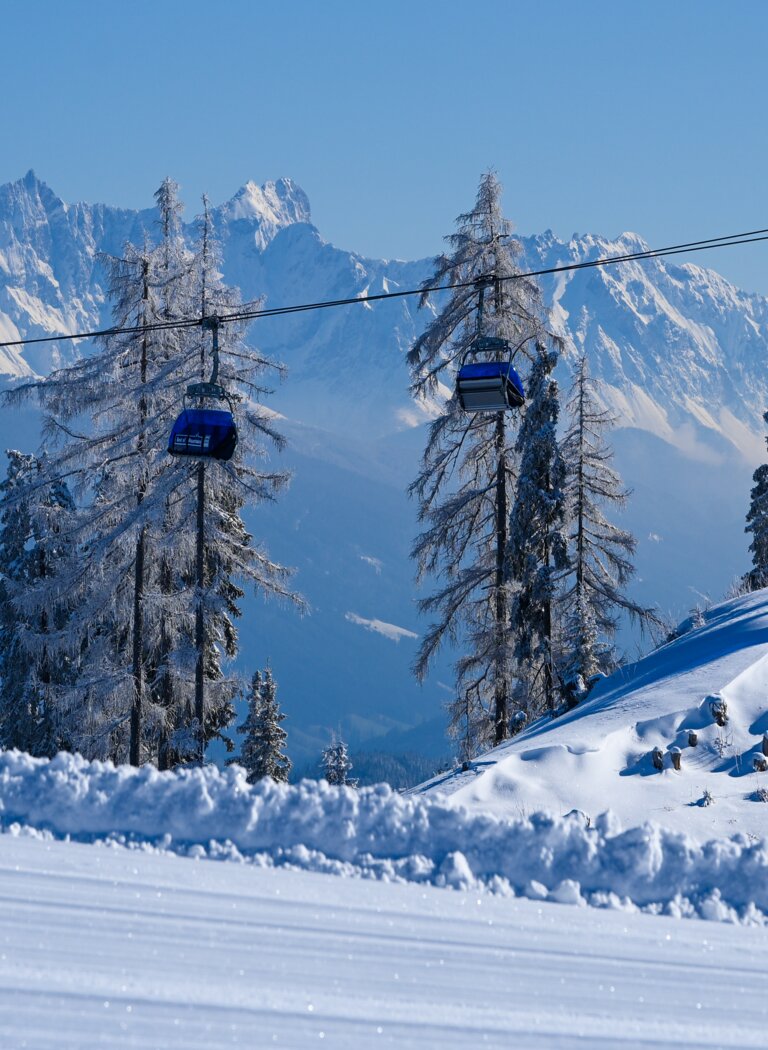 Two blue cable cars travel on a ropeway above snowy hills with frosted trees and Alps in the background | © Ski amadé GmbH