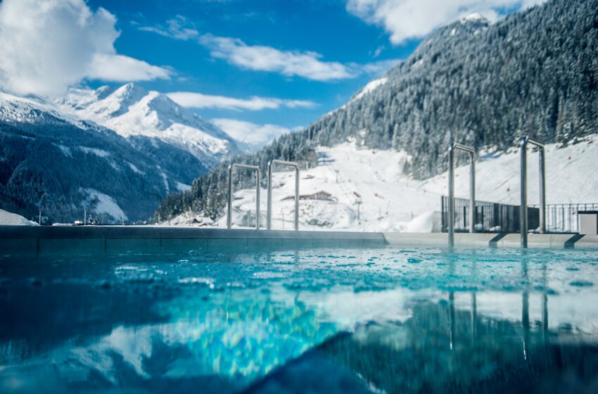 Heated outdoor pool beside snowy ski slopes and forested mountains under a blue sky in Bad Gastein | © Max Steinbauer