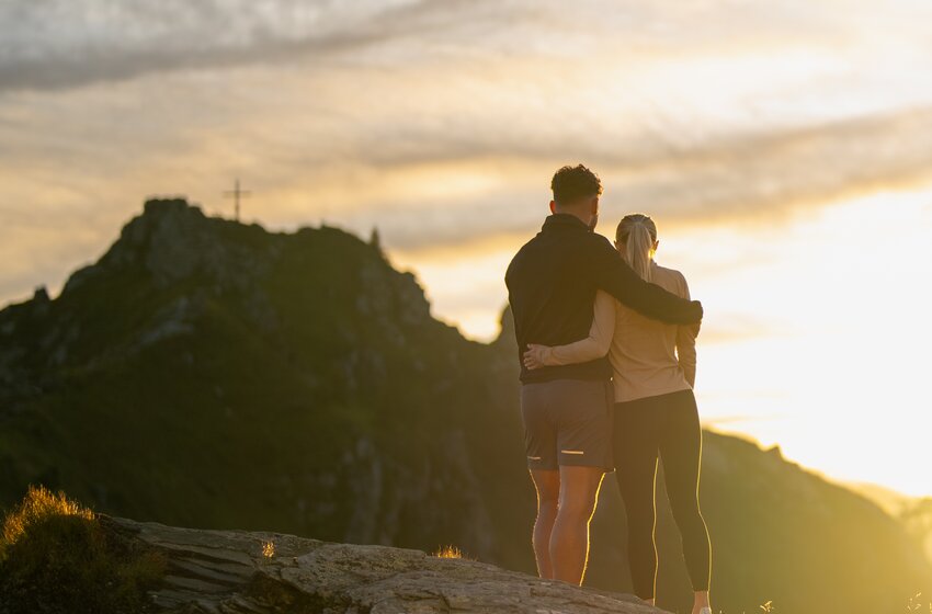 Couple stands closely embraced on rock at mountain summit at sunset, mountains and sky in background | © Gasteiner Bergbahnen AG