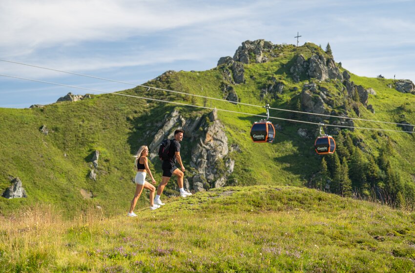 Couple hiking across green alpine meadow with backpack, gondolas behind and rocky summit with cross | © Gasteiner Bergbahnen AG