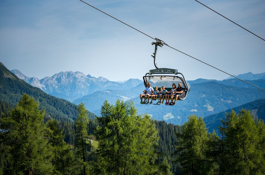 Six people ride the 6-seater Gamskogel chairlift uphill in summer, gliding above green forest with panoramic mountain views. | © Zauchensee Liftgesellschaft Benedikt Scheffer GmbH