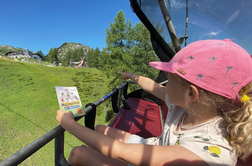Girl with pink cap holds MeisterCup card and points at adventure playground while riding downhill in the Gamskogel chairlift. | © Zauchensee Liftgesellschaft Benedikt Scheffer GmbH