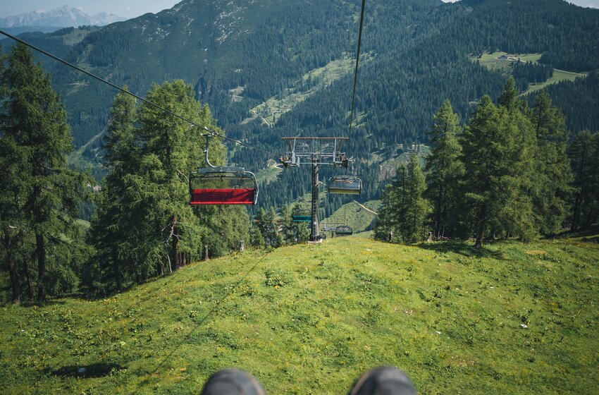 Chairlift descent with hiking boots in front, green alpine meadows below and forested mountains in the background. | © Zauchensee Liftgesellschaft Benedikt Scheffer GmbH