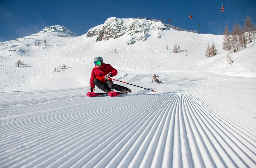 Two skiers carve turns on a groomed slope in front of snow-covered mountains with a gondola lift in sunshine. | © Zauchensee Liftgesellschaft Benedikt Scheffer GmbH