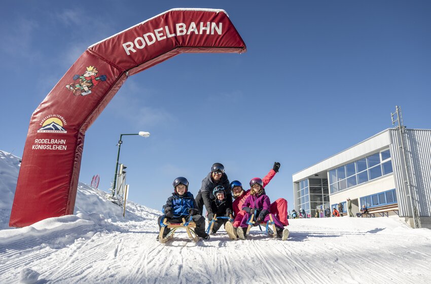 Four people on sledges under start arch of Königslehen toboggan run next to modern mountain station in sun | © Zauchensee Liftgesellschaft Benedikt Scheffer GmbH