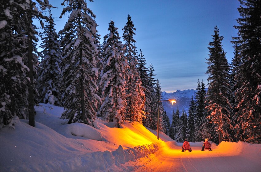 Two sledders ride at dusk on a lit snowy track through a snowy forest with mountain view | © Zauchensee Liftgesellschaft Benedikt Scheffer GmbH